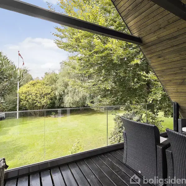 Terrasse med sorte møbler og kurv. Udsigt over en velplejet grøn have med træer og flagstang. Glaspaneler indrammer området. En lanterne hænger ned fra loftet.