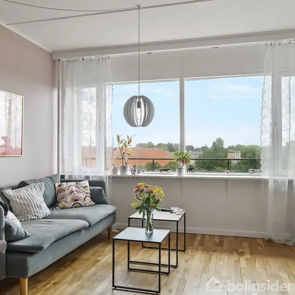 Stue med grå sofa fyldt med puder, ved siden af vindue med gardiner. Der står farverige blomster på sorte borde. Fladskærms-TV på kommode. Udsigt til træer og bygninger udenfor.
