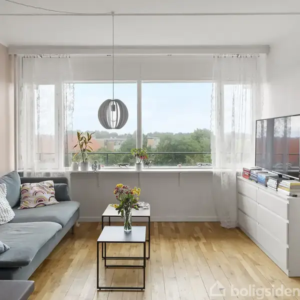 Stue med en blå sofa ved et sofabord, blomster i vaser på bord og vindueskarm. Stor vindueskarm med udsigt til grønt. Fladskærms-TV på hvid kommode.