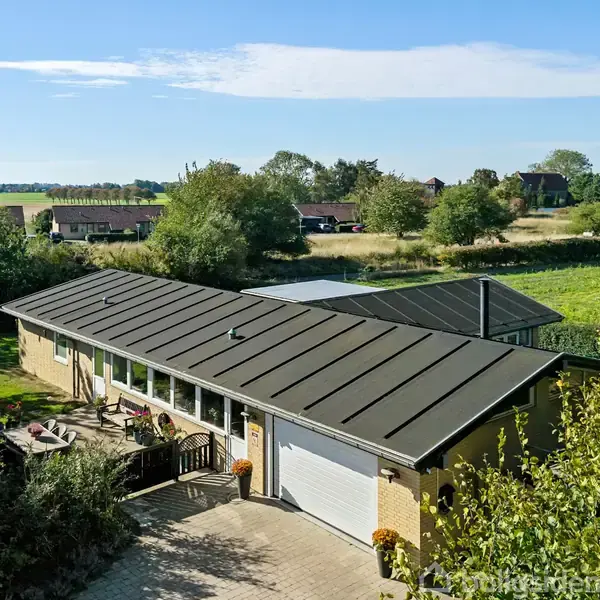 En etplans gult murstenshus med fladt tag og en terrasse foran. Det ligger i et landligt område omgivet af træer, marker og flere bygninger i baggrunden.