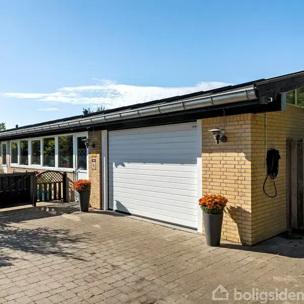 Et moderne, fladt taghus med gul murstensfacade, stor hvid garageport og blomsterkrukker. Huset er placeret i en have med flisebelagt indkørsel og omgivet af buske og træer.