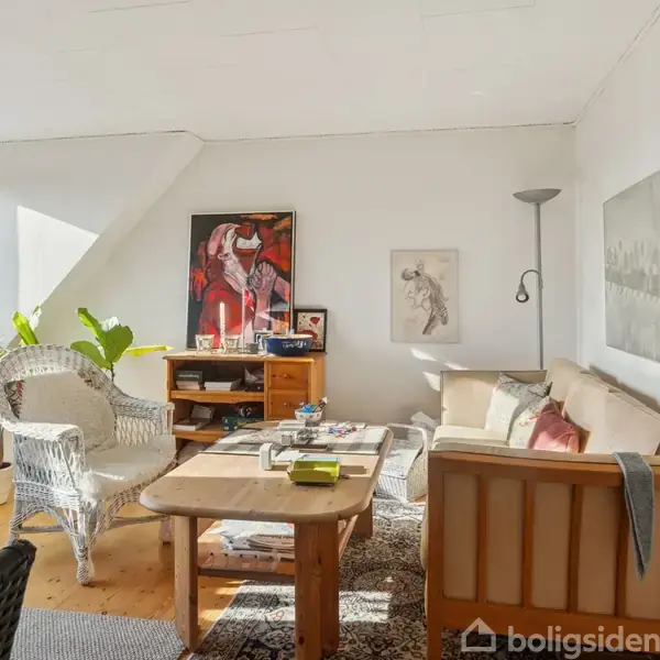 Et lyst stueområde med en sofa og lænestol omkring et træbord. Sollys strømmer gennem et vindue. Kunstværker hænger på væggene, og der er planter ved siden af.