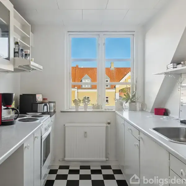 Et lyst køkken med sorte og hvide ternede gulve indeholder en kaffemaskine, ovn og vask. Vindue med udsigt til bygninger med orange tage. Planter står på vindueskarmen.