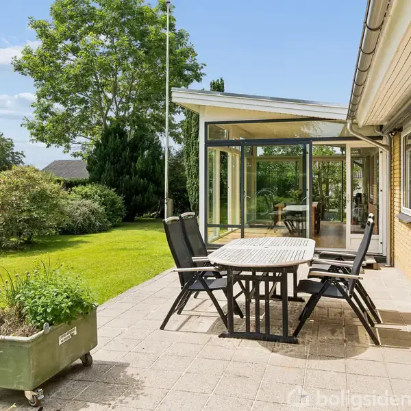 Terrassemøbler står omkring et bord på en fliset terrasse. En grøn have med træer og buske omgiver terrassen. En vinterhave ses i forlængelse af huset. Tekst på plantekasse: "Urtehave".