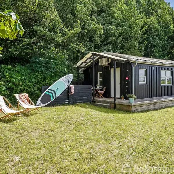 Et sort, lille sommerhus står på en græsplæne omgivet af træer. To strandstole og et surfbræt står foran huset, som har en terrasse og en plantekrukke.