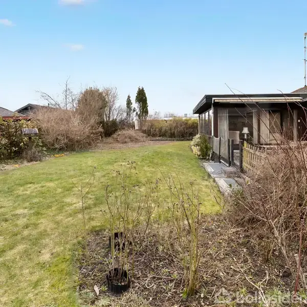 En græsplæne strækker sig langs et rækkehus, hvor en terrasse med glasparti er synlig. Omgivet af lav vegetation og buske, med flere huse i baggrunden under en blå himmel.