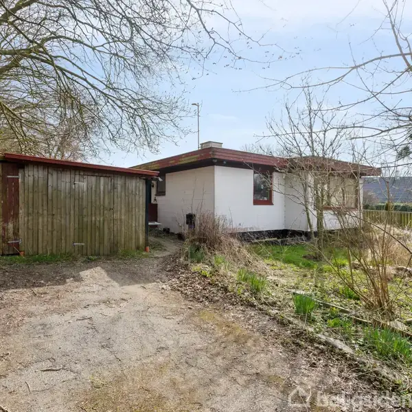 Et lille hvidt hus med fladt tag står omgivet af træer og buske. Ved siden af er der et træskur. Grussti fører til huset gennem den tilgroede have.