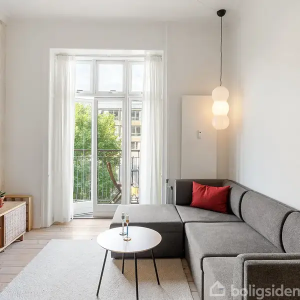 Grå hjørnesofa med rød pude står i en stue foran et lille rundt bord. En moderne lampet hænger over sofaen. I baggrunden ses balkon med åbne døre og grønne træer.
