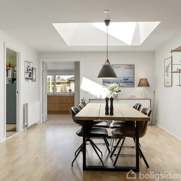En spisestue med et træbord og seks stole står under en loftslampe. I baggrunden er der adgang til et køkken. Rummet har lyse vægge og trægulv.
