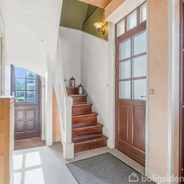 En lys gang med trappe med røde trin, der fører opad. Døren til venstre er åben og slipper sollys ind. To lukkede døre til højre. En lanterne står på trappen.