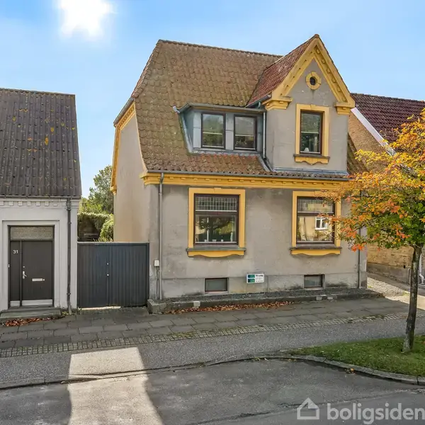 Et gråt hus med gule detaljer står på en stille villavej. Det har to etager og en brun tagkonstruktion. Ved siden af er et træ med gyldne efterårsblade.