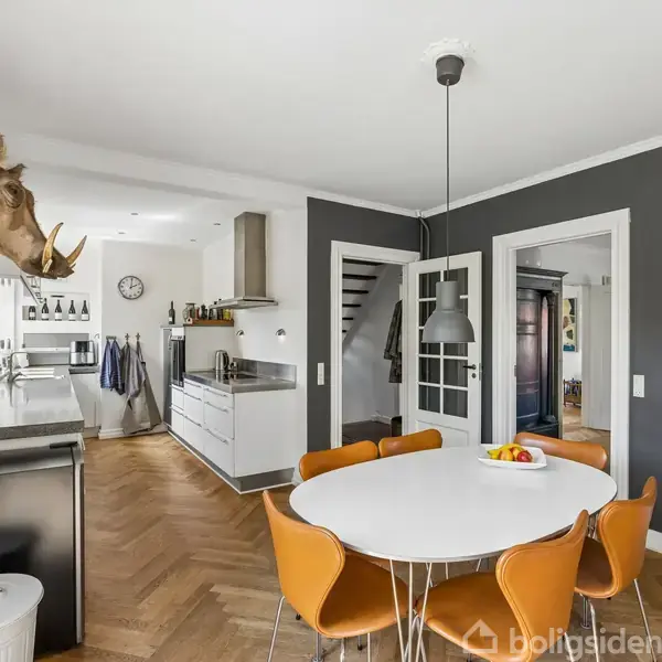 Rundt spisebord med seks brune stole står i en moderne spisestue-adskilt køkkenområde. Et næsehornshoved er monteret på væggen. Farverigt maleri pryder væggen.