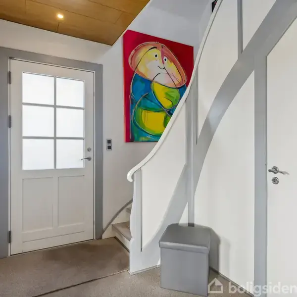 En trappegang indeholder en kurvet trappe opad, omgivet af hvide vægge. En maleri med abstrake figurer hænger ved siden af døren. Knagerække til venstre.