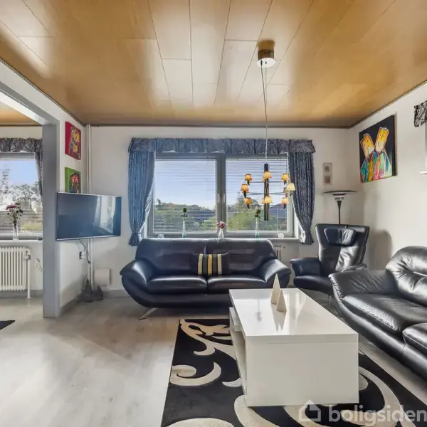 Sort lædermøblement med sofaer og stol, står i en stue. Bord i midten, hængende lysarmatur over. Ved siden af hænger kunst på væggen. Store vinduer med blå gardiner.