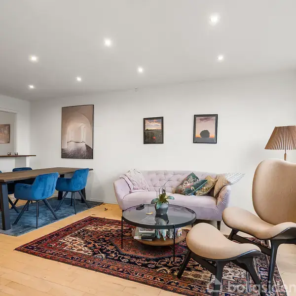 En stue indeholder en lyserød sofa, to stole og et sofabord på et tæppe. I baggrunden står et spisebord med blå stole ved siden af en reol. Ingen tekst tilstede.