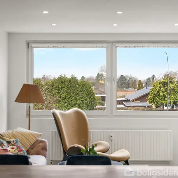 En læderstol står ved et stort vindue, der viser en udsigt over en forstadshave med træer og huse. Ved siden af stolen er en gulvlampe og en sofa.