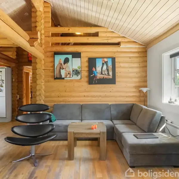 Grå hjørnesofa med sofabord i en stue af træ. Sort designerstol placeret ved siden af. Væggen har farverige fotografier, og vindue lader naturligt lys ind i rummet.
