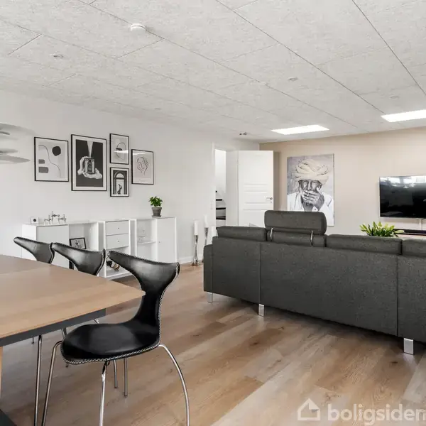Sort sofa står foran et væghængt TV i en moderne stue. Bagved ses spisebord med sorte stole og vægkunst. Rummet er lyst med trægulv og indbydende atmosfære.