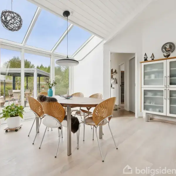 Et spisebord står midt i et lyst rum med seks stole omkring. Bordet er placeret nær store glasvægge, der åbner til en terrasse. Skab med pynt ovenpå i baggrunden.