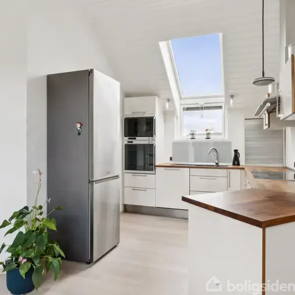 Køkken med moderne, hvidt interiør, der centrerer omkring et højt køleskab ved siden af en ovn. En grøn plante står i et hjørne, mens et farverigt billede hænger på væggen.