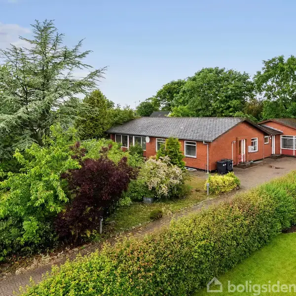 Rødt hus omgivet af frodig have med forskellige buske og træer. Asfalteret sti leder til indgangen. Skyfri himmel i baggrunden, som skaber en rolig atmosfære.