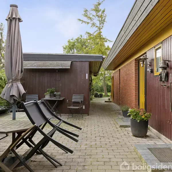 En terrasse med et bord og stol i sort træ står under en lukket parasol. Flisebelægningen strækker sig mellem røde murstensvægge og grønne buske i baghaven.