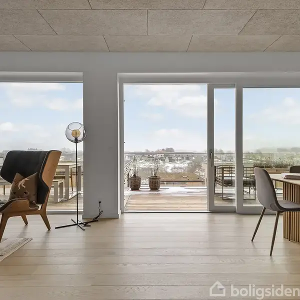 Stue med lænestol og spisebord placeret ved store vinduer. Lys strømmer ind, og en balkon med udsigt over det åbne landskab ses udenfor.