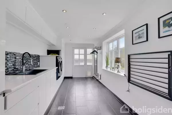 Et moderne bryggers har hvide skabe og sorte fliser. En vaskemaskine og en vask er til stede. Store vinduer med udsigt, planter og kunst på væggen.