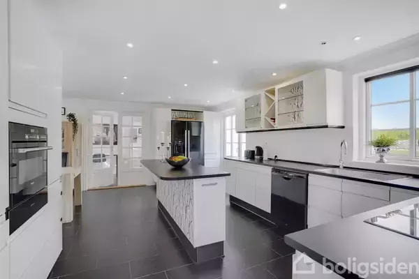 Et moderne køkken har en stor, central køkkenø. Køkkenet er lyst og rummeligt med hvide skabe og sorte bordplader. Store vinduer giver naturligt lys og udsigt til det ydre miljø.