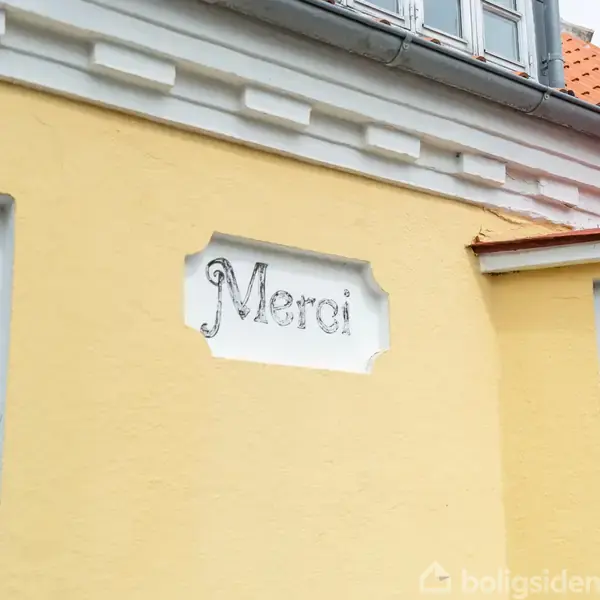 Et skilt med teksten "Merci" er indlejret i en gul væg. Skiltet har dekorative kanter, og væggen er en del af en bygning med hvidt tagudspring.
