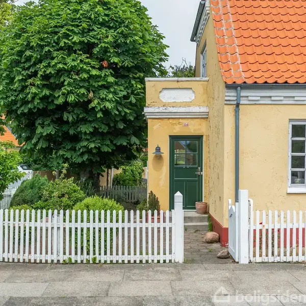 En gul bygning med grøn dør markeret med tallet "14", omgivet af en hvid stakit og have med buske og et træ i en hyggelig landsbyatmosfære.
