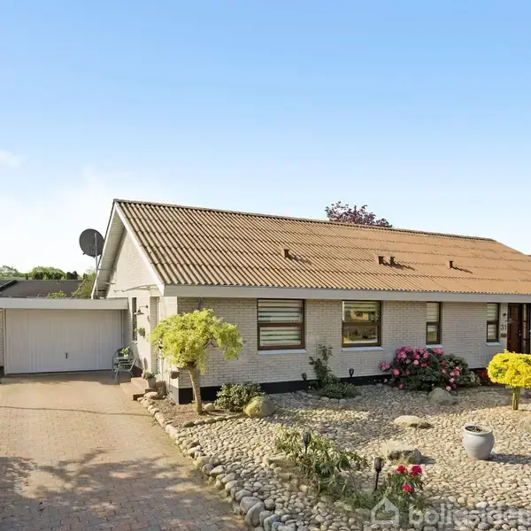 Et enkelt enfamiliehus med et tegltag, omgivet af en velholdt have med grus og dekorative planter, beliggende på en stille villavej med en garage ved siden af indkørslen.