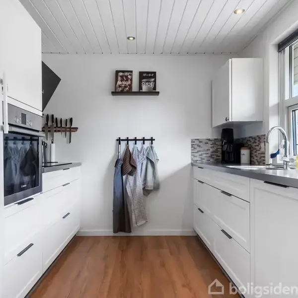 Moderne køkken med hvide skabe, indbygget ovn, og knagerække med klæder. Vinduet til højre giver udsyn til have. Loftet er hvidt med spotlys, og gulvet er i træ.