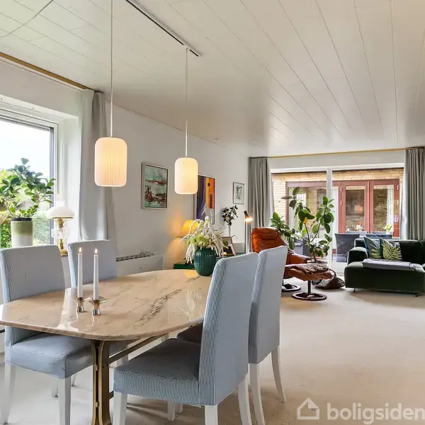 Spisebord med marmorbordplade står i en moderne opholdsstue. To pendellamper hænger over bordet, omgivet af grønne planter og moderne møbler. Store vinduer bringer lys ind i rummet.