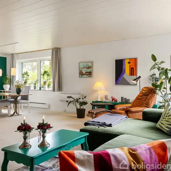 Stue med moderne indretning; sofabord med blomster og lys, stor hjørnesofa med farverige puder; spiseområde med spisebord og stole ved vindue; kunst og planter dekorerer rummene.