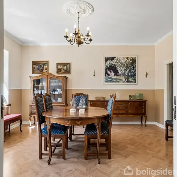 Rundt spisebord med seks stole står midt i et elegant rum. En lysekrone hænger over bordet, omgivet af klassiske møbler og kunst på væggene. Trægulv og lyse vægge dominerer rummet.