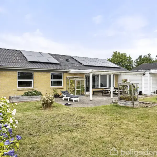 Et murstenshus med solpaneler på taget har en terrasse med liggestol og spisebord. Haven indeholder græs og blomsterbuske, samt en nedgravet trampolin.