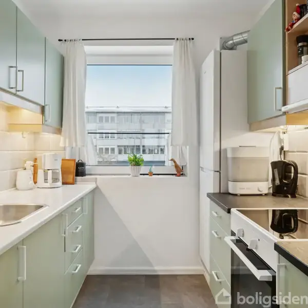 Et moderne køkken med grønne skabe, en vask og en ovn. På bordpladen står en kaffemaskine og en brødrister. Et vindue med gardiner viser en bygning udenfor.