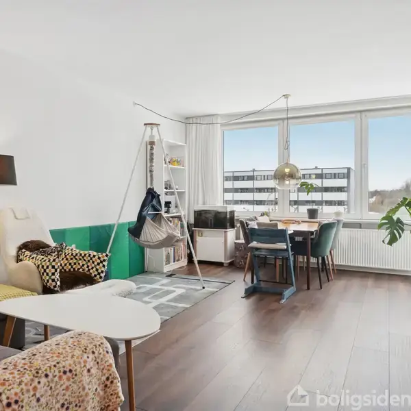 Stue med trægulv har en siddeplads med en hvid lænestol og lampe, et spisebord nær store vinduer, og grønne planter. En hængekøje er monteret i hjørnet under hylder.