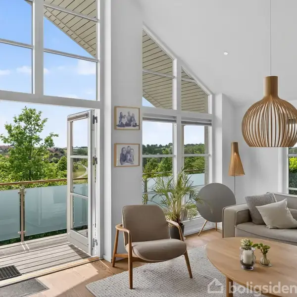 Stue med sofa, lænestol og sofabord, hvor store vinduer og glasdør står åbne ud mod en terrasse og have. En moderne lampe hænger fra det høje, lyse loft.