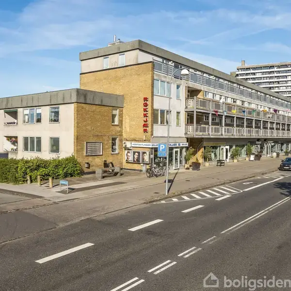 En gul murstensbygning med tre etager og en lang balkon har skiltet "RØDOVRE CENTRUM." Den ligger ved en vej med parkerede biler og grønne buske.