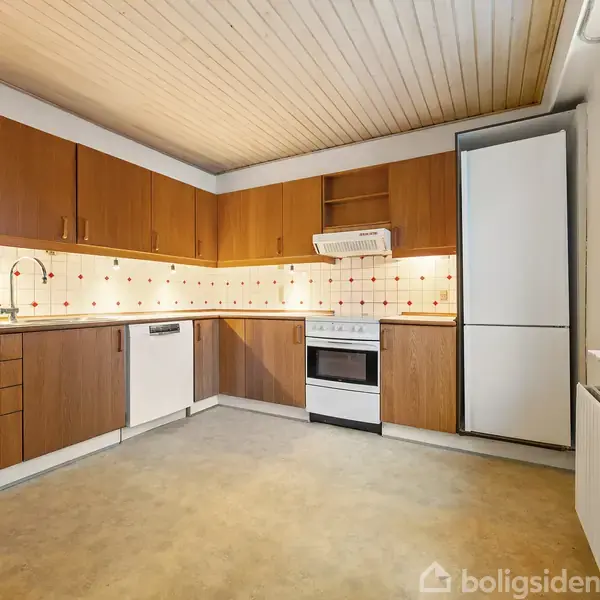 Køkken med træskabe og skuffer, arrangeret i vinkel med en opvaskemaskine, komfur og køleskab. Flisevæg med mønster og træloft. Store vinduer til højre.