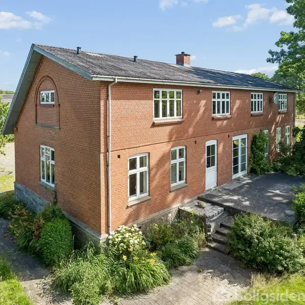 Rødt murstenshus med hvide vinduer står på en velplejet grund. Trappe fører til hoveddør. Omgivet af grønne planter og græs i et landligt område.