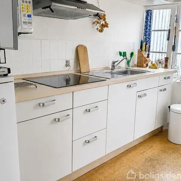 En trækommode med en lampe og billeder ovenpå står ved en vindeltrappe i et lyst rum. Ved siden af er et tøjstativ med hængende jakker. Vinduet viser udsigt udenfor.