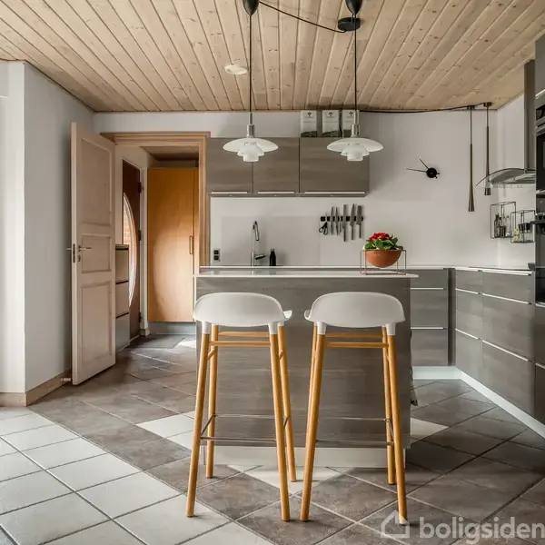 Køkken med moderne grå skabe og indbygget ovn, to høje barstole ved køkkenø. Store vinduer giver udsigt til have. Træloft og flisegulv skaber en lys og behagelig atmosfære.