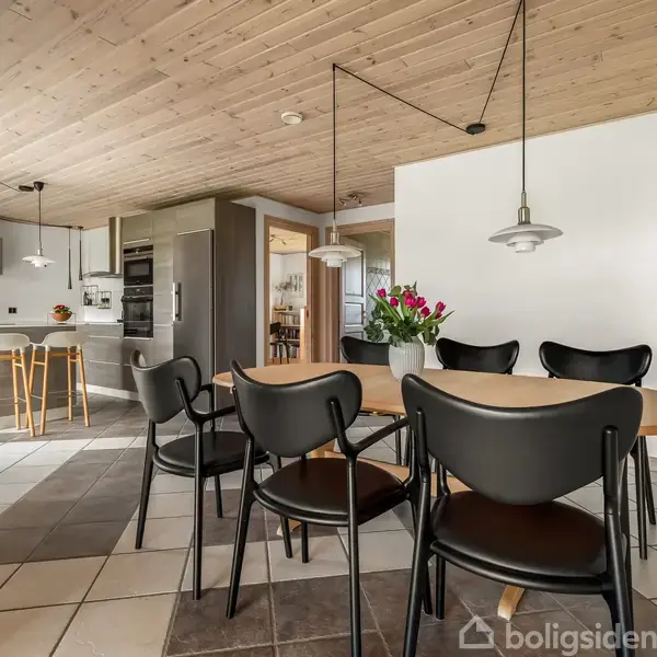 Rundt spisebord med seks sorte stole står i et køkken-alrum. I baggrunden ses et moderne køkken med barstole ved en disk. Blomster pryder både bord og køkkenbord.