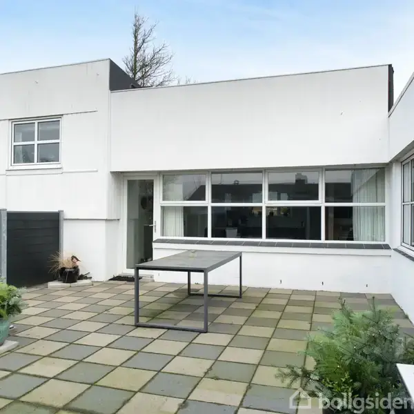 En rektangulær metalbord står på en terrasse med grå fliser. Planter i krukker placeres rundt om, og et moderne, hvidt, toetagers hus omkranser terrassen.