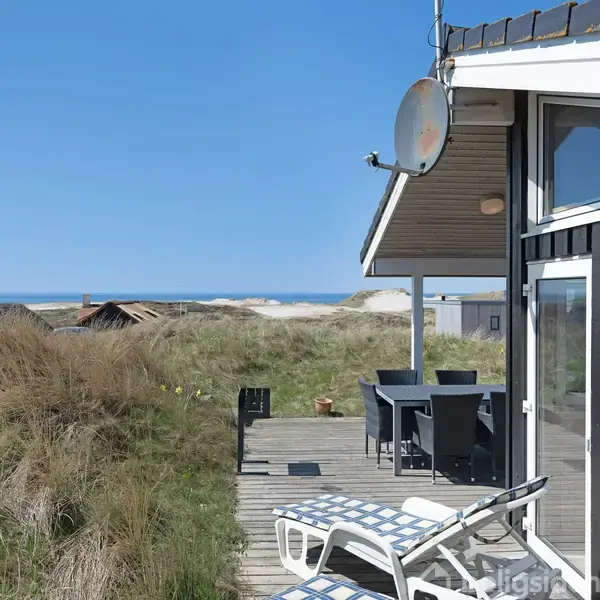 En sort træbygning med terrassebord og liggestole står på en træterrasse. I baggrunden ses klitter og hav under en klar blå himmel.