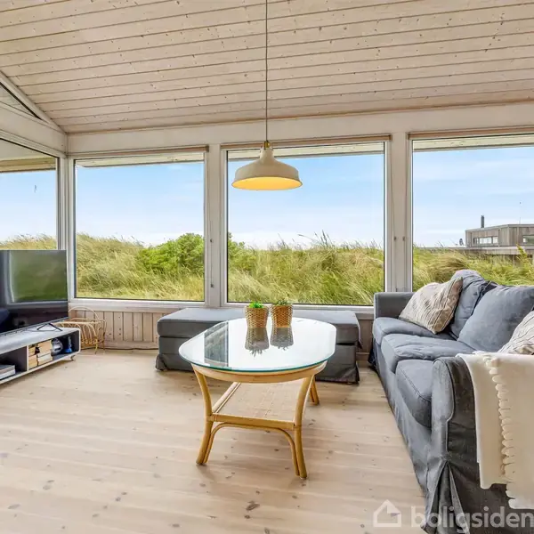 Stue med grå sofa, sofabord og fladskærms-tv. Store vinduer viser udsigt til græs og moderne hus i baggrunden. Lyse trægulve og loft skaber en rummelig atmosfære.