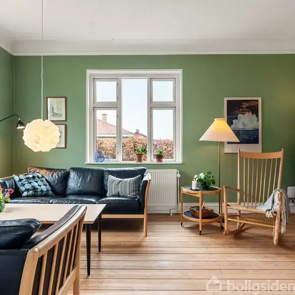 Stue med sorte lædersofaer og lænestol omkring et sofabord. Grønt malede vægge, træhylder med bøger og lamper. Vinduer med udsigt, planter i vindueskarmen, trægulv.